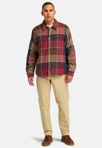 Camisa de cuadros en marrón, rojo y azul marino, con un bolsillo en el pecho y botones a presión, combinada con pantalones beige y zapatos marrones.