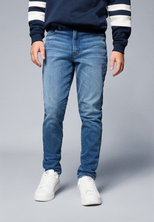 Man draagt middenblauwe slim-fit jeans, marineblauwe trui met witte strepen op de mouwen en witte lage sneakers, staand tegen een grijze achtergrond.