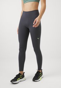 Legginsy w kolorze ciemnoszarym, o wysokim stanie, wykonane z elastycznego materiału, z małym neongrębnym logo na boku, zestawione z czarnymi butami sportowymi.