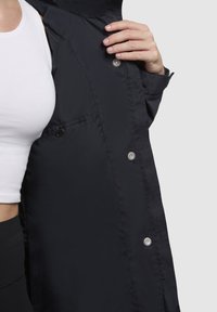 Black lightweight jacket featuring a snap closure, smooth texture, and a side pocket with a button. Accented with a high collar.