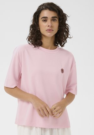 Femme aux cheveux bouclés et foncés, portant un t-shirt à manches courtes rose clair avec une petite broderie de fleur rouge foncé sur la poitrine.