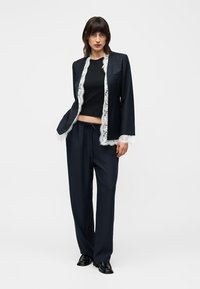 Ženska nosi črne široke hlače in ujemajoč se črni blazer z belo čipkasto obrobo, kombinirano s črnim kratkim topom in črnimi čevlji, stoječa.