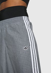 Pantalones deportivos grises con tela texturizada, con tres rayas blancas a lo largo de los lados y un logo negro bordado en el muslo izquierdo.