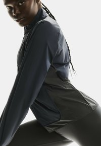Veste de sport pour femmes en marine foncé, avec un col haut, un matériau léger et des accents noirs contrastants au bas et sur les panneaux latéraux.