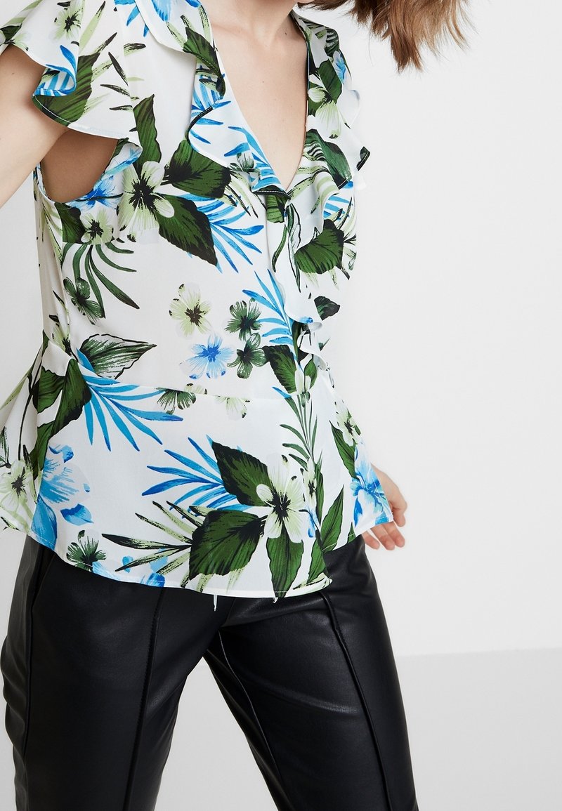 Blusa floreale bianca con foglie verdi e fiori blu, con maniche a volant e scollatura a V. Abbinata a pantaloni in pelle nera.