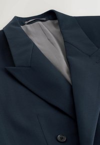 Blazer bleu marine avec revers crantés, un bouton noir unique et une doublure intérieure gris clair, posé à plat sur une surface blanche.