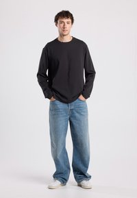 Long-sleeve black t-shirt paired with loose-fitting light blue jeans. Model wears white sneakers, standing with hands in pockets.