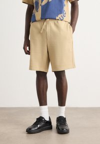 Shorts de sport beiges avec un waistband élastique, poches latérales et une coupe décontractée, associés à des baskets noires et des chaussettes blanches montantes.