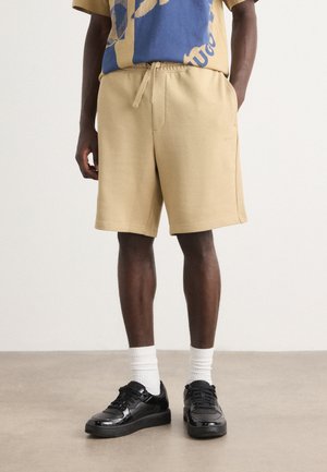 Beige sportshorts med elastisk midja, sidfickor och avslappnad passform, matchade med svarta sneakers och vita ankelsockor.