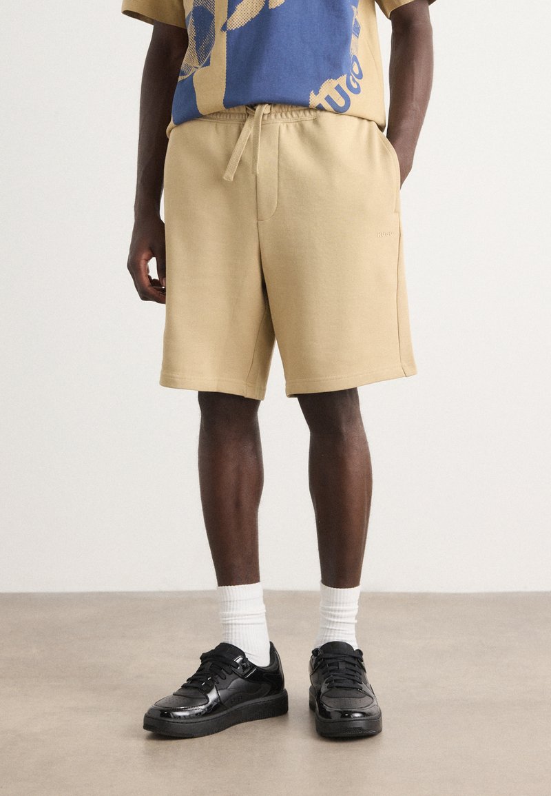 Shorts de sport beiges avec un waistband élastique, poches latérales et une coupe décontractée, associés à des baskets noires et des chaussettes blanches montantes.