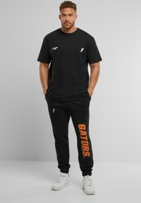 Sort t-shirt med hvidt logo og orange "GATORS" tryk på sorte sweatpants. Letvægtsstof, afslappet pasform og elastisk talje. Hvide sneakers.