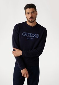 Granatowy sweatshirt wykonany z mieszanki bawełny, z teksturowanym logo "GUESS EST. 1981" w odcieniu niebieskim. Ściągacze na rękawach i u dołu, klasyczny dekolt.
