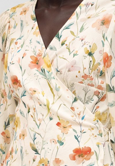 Άτομο με σκουρόχρωμο δέρμα που φοράει ένα κρεμ φόρεμα τύπου wrap με μακριά μανίκια, με πολύχρωμο, λεπτό, ανθισμένο μοτίβο σε απαλό στυλ ακουαρέλας.