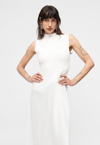 Γυναίκα με τατουάζ ποζάρει με τα χέρια στη μέση, φορώντας λευκό φόρεμα χωρίς μανίκια με ψηλό γιακά και μακριά κρεμαστά σκουλαρίκια, σε απλό φόντο.