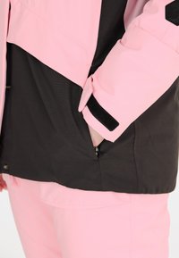 Wasserabweisende Jacke mit einem pinkfarbenen Oberteil und einem schwarzen unteren Abschnitt, ausgestattet mit einer Reißverschlusstasche und Klettverschluss an den Ärmeln.