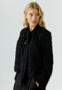Blonde Frau, die nach rechts schaut, trägt eine strukturierte schwarze Felljacke über einer schwarzen Spitzenbluse mit einem Schlipskragen.