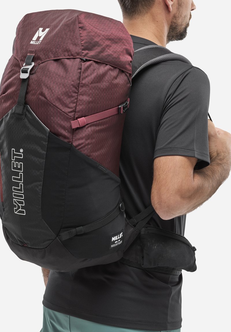 Homme portant un grand sac à dos de randonnée Millet maroon et noir et une chemise noire, vu de côté et de dos.