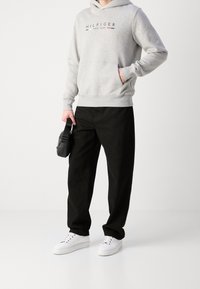 Felpa grigia in cotone con stampa del logo, tasca frontale, abbinata a pantaloni neri e sneakers bianche. Gli accessori includono una piccola borsa nera.