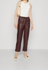 Bordowe skórzane spodnie z elastycznym pasem, zestawione z dopasowanym białym topem na ramiączkach oraz białymi butami na obcasie z złotymi akcentami.