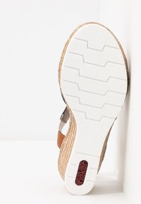 Semelle blanche de sandale compensée avec des côtés en osier marron et des sangles noires et brunes, affichant le logo de la marque 'rieker' en rouge.