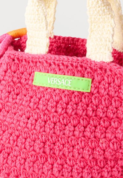 Χειροποίητη ροζ τσάντα με χοντρές κρεμ λαβές και πράσινη ετικέτα στην πρόσοψη που εμφανίζει το όνομα της μάρκας «Versace».