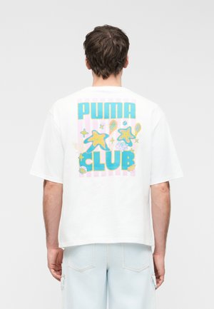 Vyras vilki baltą per didelį marškinėlį su spalvingu užrašu „PUMA CLUB“ ir nugaros pusėje esančiomis jūros žvaigždžių, kriauklių ir teniso raketės grafinėmis detalėmis.