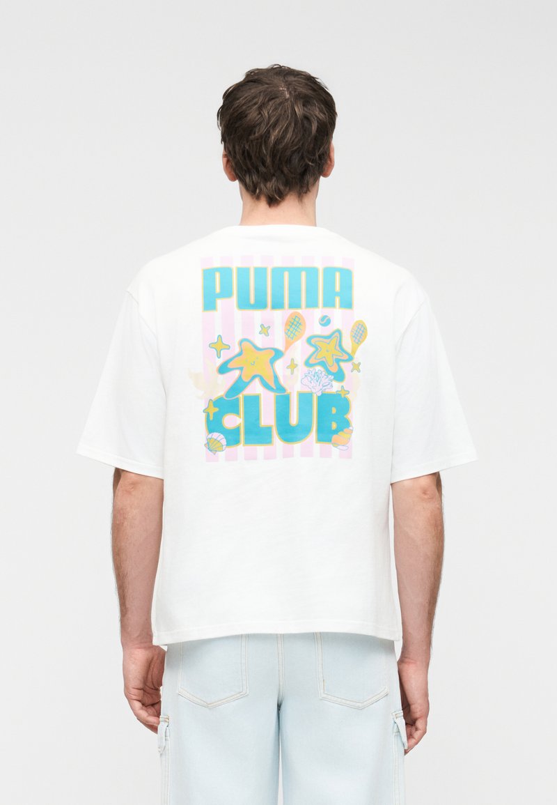 Άνδρας που φοράει λευκό υπερμεγέθες T-shirt με πολύχρωμο κείμενο "PUMA CLUB" και γραφικά αστερία, κοχυλιών και ρακέτας τένις στην πλάτη.