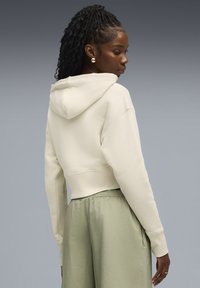 Beige cropped hoodie met een capuchon, ontspannen pasvorm mouwen en ribgebreide afwerking. Model draagt saliegroene broeken die een gladde stof en elastische taille tonen.