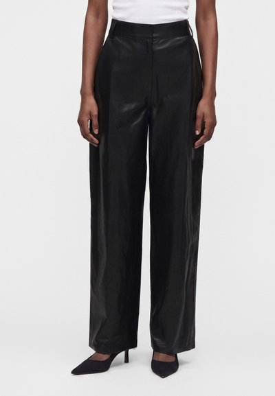Personne portant un pantalon en cuir noir taille haute et à jambes larges, des talons noirs à bout pointu et un haut blanc visible à la taille.