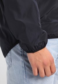 Svart lättviktsjacka med elastiska ärmslut, med en slät textur och rak nederkant, matchad med ljusblå denimjeans.