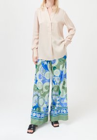 Vrouw in beige blouse en wijde, gestreepte broek in groen, blauw en wit met zwarte open-toe sandalen, staat tegen een witte achtergrond.