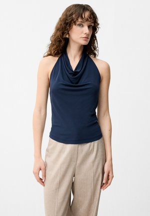 Femme aux cheveux bouclés portant un haut bleu marine sans manches à col bénitier et un pantalon beige à jambes larges, debout devant un fond blanc uni.