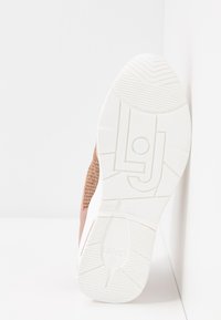 Semelle blanche d'une basket rose avec une bande texturée et le logo "LJ" en relief, inclinée contre un mur blanc sur une surface blanche.