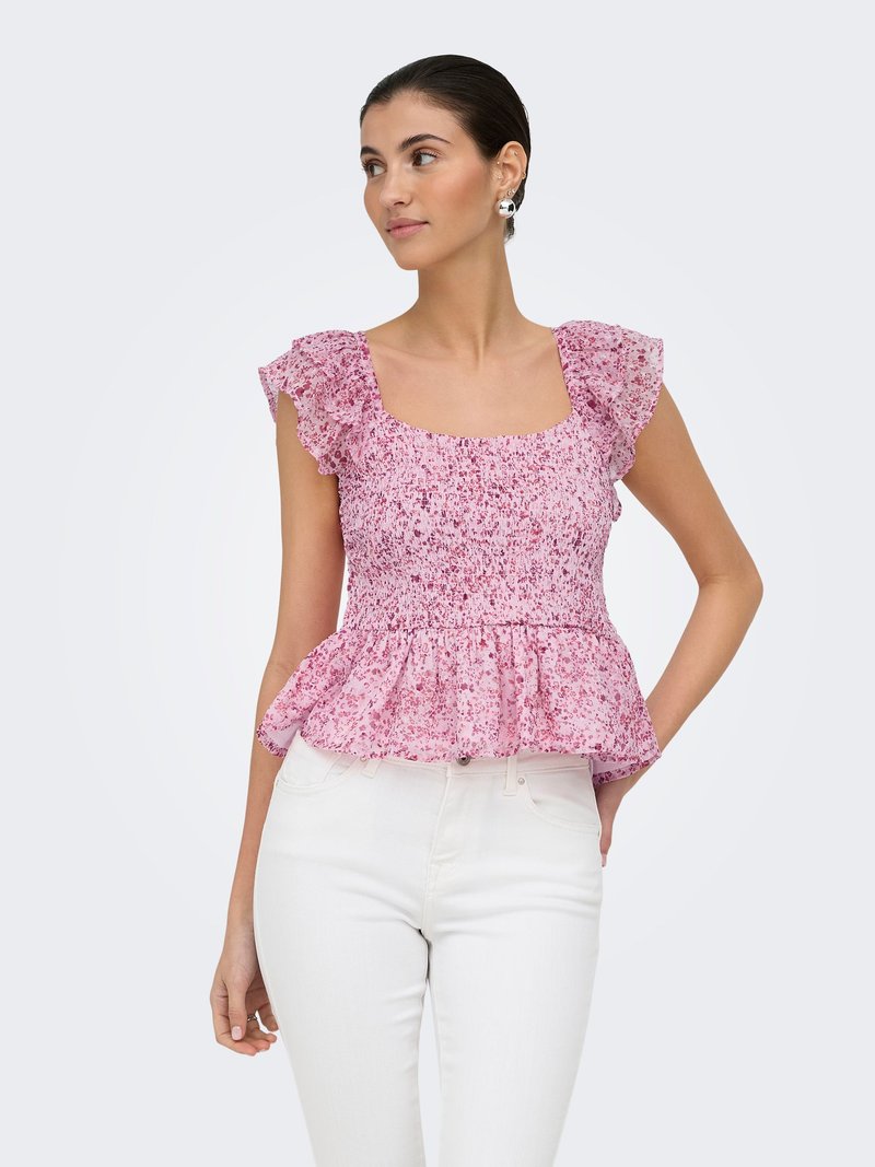 Femme portant un haut rose à volants avec un motif floral et un pantalon blanc, debout, une main dans la poche, regardant sur le côté.