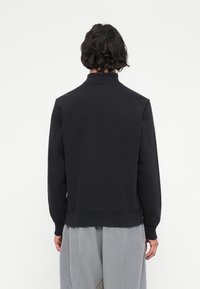 Άτομο με κοντά σγουρά μαλλιά που φοράει ένα μαύρο μπλουζάκι με μακριά μανίκια και ανοιχτό γκρι παντελόνι φλις, γυρισμένο μακριά απέναντι σε ένα λευκό φόντο.