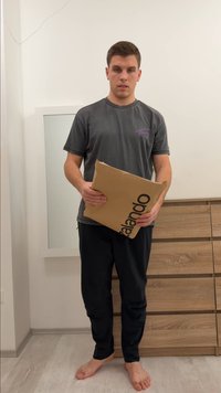 Rjavo kartonsko pakiranje s črnim tiskom "Zalando", držano pokonci. V ozadju lesena komoda in ogledalo. Oseba nosi sivo majico in temne hlače.