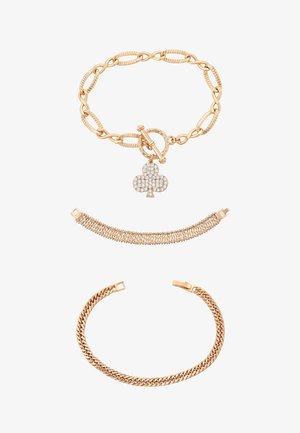 Trzy złote bransoletki: jedna z dużymi, teksturowanymi ogniwami i zawieszką w kształcie pałki wysadzaną diamentami, jedna o splatanym wzorze oraz jedna z gładkim łańcuszowym wzorem.