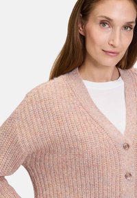 Hellrosa gestrickte Strickjacke mit V-Ausschnitt, geripptem Muster und zwei Knöpfen auf der Vorderseite. Über einem weißen Oberteil getragen.