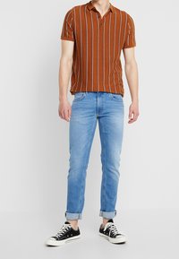 Brunrandig polo skjorta med korta ärmar, tillsammans med ljusblå jeans med uppvikta ärmar och svarta sneakers med vita sulor.