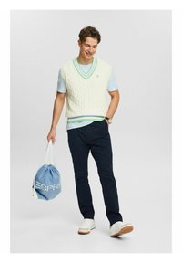 Pletený krémový svetřík bez rukávů se zeleným lemováním, pod ním světle modrá košile, tmavě modré kalhoty a bílé tenisky, držící modrý stahovací batoh.