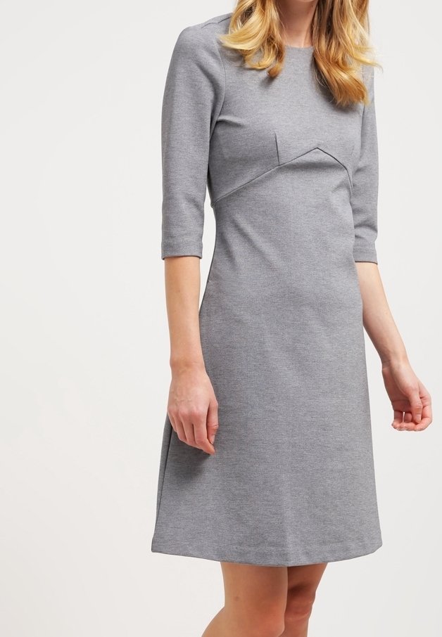 Femme portant une robe grise cintrée, longueur genou, avec manches trois-quarts et des détails de couture subtils sur la poitrine.