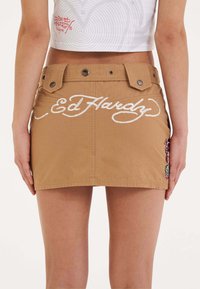 Ruskean farkkuminihame, jossa on valkoiseen brodeerattuun "Ed Hardy" -logoon ja koristeelliseen brodeeraukseen sivuilla. Kaksi näkyvää nappikoristetta.
