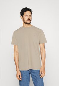 T-shirt beige a maniche corte realizzato in tessuto morbido, con collo rotondo e un sottile dettaglio del logo sul petto, abbinato a jeans in denim blu.