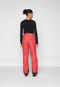 Pantalons de ski rouges en tissu durable, avec une taille haute, des poches latérales et un bas cintré. Associés à un t-shirt noir à manches longues.