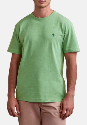 Homme portant un t-shirt à manches courtes vert clair moucheté avec un petit logo d'arbre vert foncé sur la poitrine gauche et un pantalon beige.