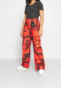 Röda och bruna mönstrade byxor med vid ben och hög midja, matchade med en svart kortärmad topp och vita skor. Liten väska fäst vid midjebandet.