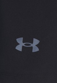Logo de Under Armour reflectante centrado en tela negra texturizada, con costura visible en el borde inferior.