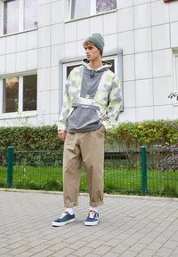 Pullover cinza claro e verde com padrão, com um bolso frontal, combinado com calças largas bege e ténis multicoloridos, ao ar livre.
