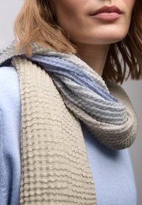 Tekstureret tørklæde med bølgende mønstre i lys beige, blød blå og dæmpet grå, afslappet draperet om halsen mod en blå top.