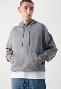 Siv hoodie z sprednjo žepom, kapuco z vrvico in rebrastimi manšetami. Ima strukturirano nalepko na prsih. Obučen čez belo majico.